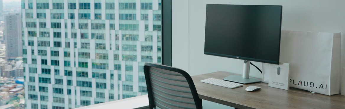 Moderner Büroarbeitsplatz mit Monitor, weißer Plaud.ai Tasche und einer Plaud Note Box vor einem Hochhausfenster.