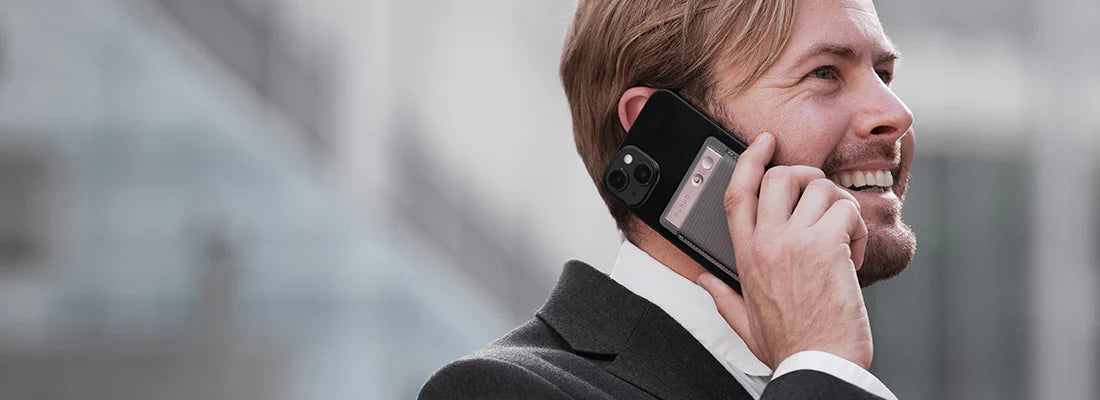 Ein lächelnder Mann im Anzug telefoniert mit einem Smartphone und einem silbernen Plaud Note auf der Rückseite.