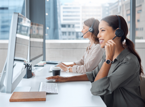 Zwei lächelnde Mitarbeiterinnen mit Headsets arbeiten an Computer-Arbeitsplätzen in einem modernen Büro.